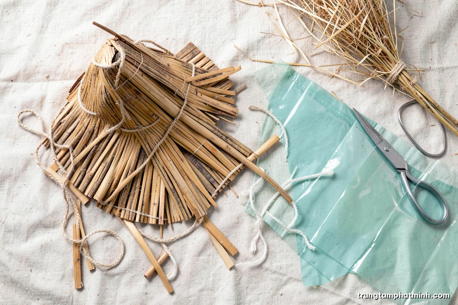 Top-down flat lay of a disassembled conical palm leaf hat beside bamboo strips, cotton cord, recycled plastic sheets and scissors on linen, soft diffused light, straw and sage palette